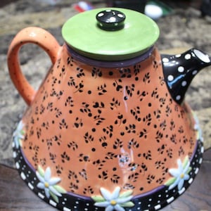 Puede incluir: Una tetera de cerámica decorativa con un cuerpo naranja vibrante adornado con patrones de hojas y puntos negros. Cuenta con una tapa verde con lunares negros, un pico negro con lunares blancos y un asa. La base está decorada con flores blancas y un borde blanco y negro.