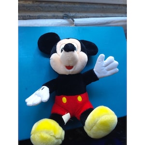 Może przedstawiać: Pluszowa zabawka Mickey Mouse z czarnym futerkiem, czerwonymi spodenkami i ż&oacute;łtymi butami. Zabawka siedzi na niebieskiej powierzchni i ma przyjazny wyraz twarzy.