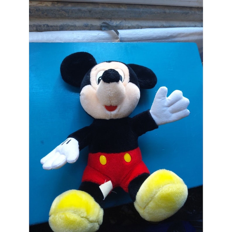 Może przedstawiać: Pluszowa zabawka Mickey Mouse z czarnym futerkiem, czerwonymi spodenkami i ż&oacute;łtymi butami. Zabawka siedzi na niebieskiej powierzchni i ma przyjazny wyraz twarzy.