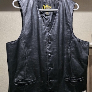Puede incluir: Chaleco de cuero negro con cuello en V y cierre de botones. El chaleco tiene dos bolsillos delanteros y está colgado de una percha blanca. La etiqueta Adler Leather Mfg. Co. es visible.