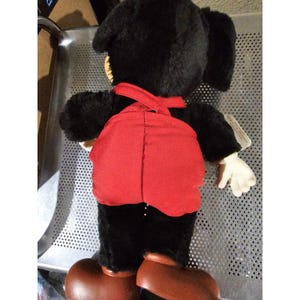 Pu&ograve; includere: Giocattolo di peluche nero con una salopette rossa. Il giocattolo ha grandi orecchie e un viso amichevole.