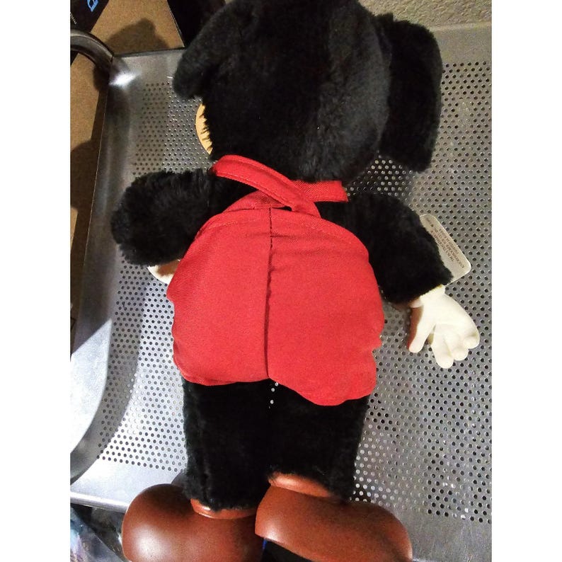 Pu&ograve; includere: Giocattolo di peluche nero con una salopette rossa. Il giocattolo ha grandi orecchie e un viso amichevole.