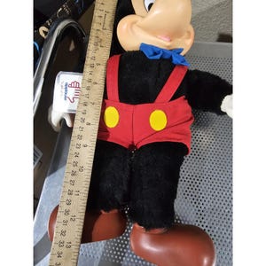 Pu&ograve; includere: Un giocattolo di peluche Mickey Mouse nero con pantaloncini rossi, bottoni gialli e un papillon blu. Il giocattolo &egrave; in piedi su una superficie bianca con un righello sullo sfondo.