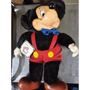 Pu&ograve; includere: Un giocattolo di peluche di Topolino che indossa pantaloncini rossi con bottoni gialli e bretelle blu. Il giocattolo ha un papillon blu e un'etichetta bianca che dice "Applause" con un'icona di mano.