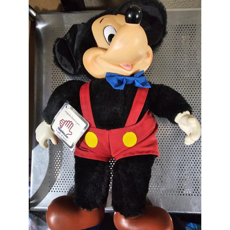 Pu&ograve; includere: Un giocattolo di peluche di Topolino che indossa pantaloncini rossi con bottoni gialli e bretelle blu. Il giocattolo ha un papillon blu e un'etichetta bianca che dice "Applause" con un'icona di mano.