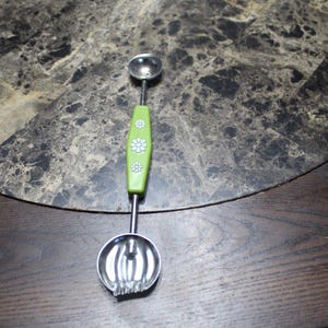 May include: A dual-ended stainless steel melon baller with a green handle. The handle is decorated with white daisy-like flowers. One end is a small scoop, and the other is a larger, fluted scoop.