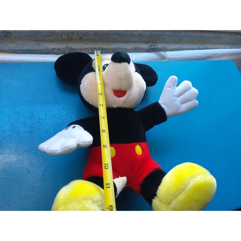 Może przedstawiać: Pluszowa zabawka Mickey Mouse z czarnymi uszami, czarnym ciałem, czerwonymi spodenkami z ż&oacute;łtymi guzikami i ż&oacute;łtymi butami. Zabawka siedzi na niebieskiej powierzchni i ma około 28 cm wysokości.