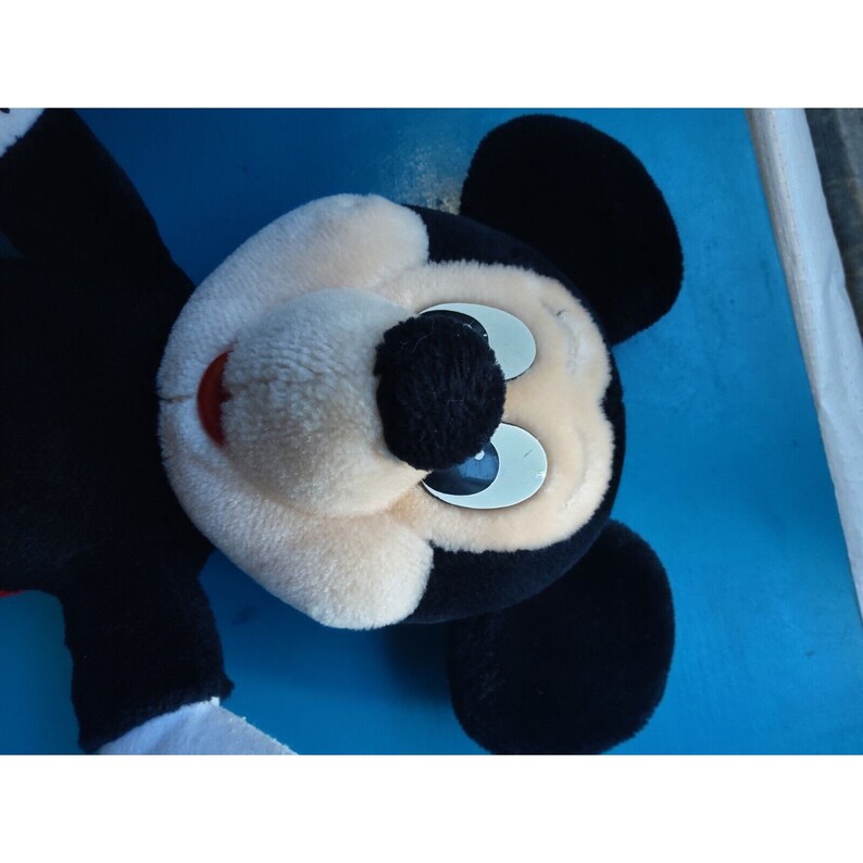 Może przedstawiać: Pluszowa zabawka Mickey Mouse w kolorze czarnym i białym z dużymi, białymi oczami i czerwonymi ustami. Zabawka leży na niebieskiej powierzchni.