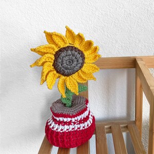 May include: A crocheted sunflower with a yellow centre and brown petals in a red and white striped pot.