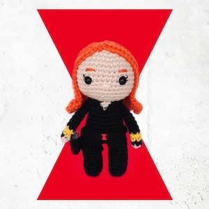 May include: A crocheted Black Widow doll with orange hair, black clothing, and a black widow symbol on her belt. The doll is standing on a red background with a white triangle.