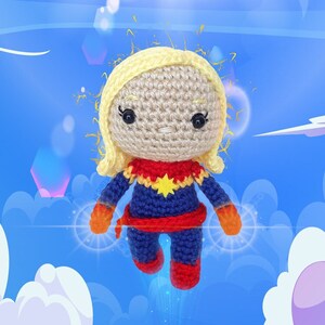 Puede incluir: Una muñeca de amigurumi de crochet de Captain Marvel, una superheroína del universo Marvel Comics. La muñeca lleva un traje rojo, azul y amarillo con una estrella en el pecho. La muñeca tiene el pelo rubio y una expresión seria.