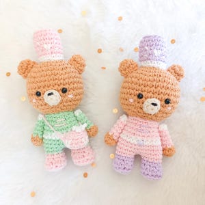 Puede incluir: Dos osos de peluche tejidos a crochet que llevan sombreros y atuendos. Un oso lleva un atuendo verde con un sombrero rosa y el otro oso lleva un atuendo rosa con un sombrero morado.