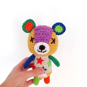 May include: A crocheted stuffed animal resembling a bear with a purple head, yellow body, and colorful patchwork clothing. The bear has a white face with black eyes and a brown nose. The bear is wearing a white shirt with red and yellow stars and has green, blue, and orange legs.