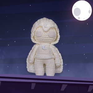 Puede incluir: Juguete de peluche blanco tejido a crochet con un diseño de media luna en el pecho y la capucha. El juguete está de pie en una repisa morada sobre un fondo azul oscuro con estrellas blancas.