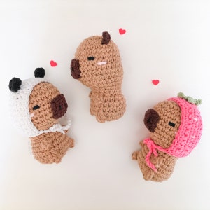 May include: Three crocheted stuffed animals in the shape of pugs, each wearing a different hat. One pug is wearing a white and black panda hat, another is wearing a pink strawberry hat, and the third is wearing a brown hat.