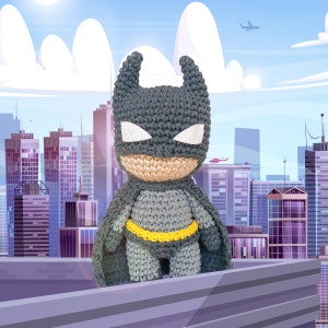 Puede incluir: Un juguete de Batman de crochet gris con un cinturón amarillo de pie en una azotea contra un horizonte de ciudad.