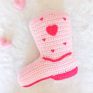 May include: Pink crocheted boot shaped pillow with red heart accents. The pillow has a pink crocheted heart in the center and smaller red hearts around the boot.