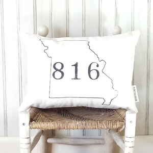 May include: White pillow with a black outline of the state of Missouri and the number 816 printed on it. The pillow is sitting on a white wooden stool with a woven seat.