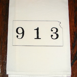 May include: White kitchen towel with a black outline of the state of Kansas and the numbers 913 printed inside.