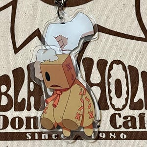 May include: A clear acrylic keychain featuring a cartoon character with a box-shaped head and a tan body. The character has a sad expression and is wearing a red bow. The keychain is hanging in front of a canvas bag with the words "BLA HOLE Cafe Since 1986".