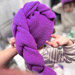 Può includere: Un cappello di lana viola brillante con una lunga coda intrecciata. Il cappello è realizzato con un filato spesso e strutturato e sembra fatto a mano. Il cappello è tenuto da una persona. Un'altra persona indossa un cappello simile sullo sfondo.
