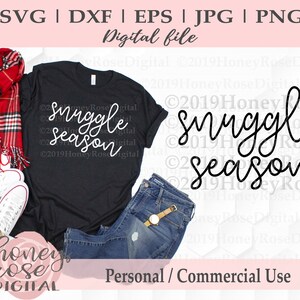Puede incluir: Una camiseta negra con el texto "Snuggle Season" en letras blancas. La camiseta se muestra sobre un fondo blanco con un par de vaqueros, una bufanda de cuadros rojos y zapatillas blancas.