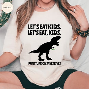 Puede incluir: Una camiseta blanca con una silueta negra de un Tyrannosaurus Rex comiendo un palo verde. El texto "Let's eat kids. Let's eat, kids." est&aacute; impreso en negro sobre el dinosaurio. El texto "Punctuation saves lives" est&aacute; impreso en negro debajo del dinosaurio.