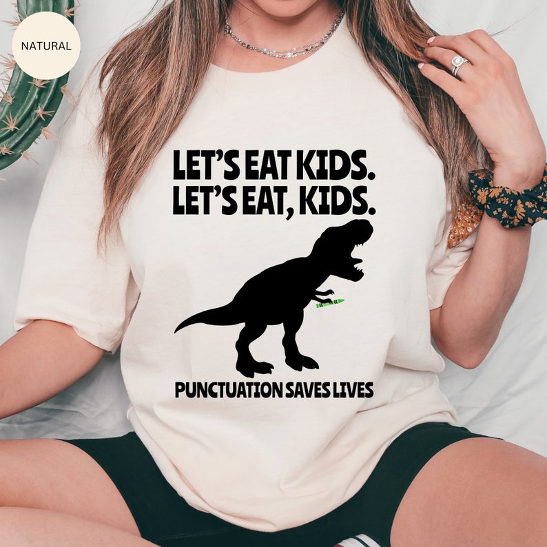 Puede incluir: Una camiseta blanca con una silueta negra de un Tyrannosaurus Rex comiendo un palo verde. El texto "Let's eat kids. Let's eat, kids." est&aacute; impreso en negro sobre el dinosaurio. El texto "Punctuation saves lives" est&aacute; impreso en negro debajo del dinosaurio.