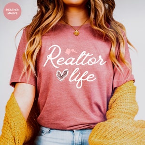 May include: A heather mauve t-shirt with the text "Realtor life" in white script. A pink heart with a leopard print design is next to the word "life".