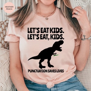 Puede incluir: Una camiseta rosa con un gr&aacute;fico de dinosaurio negro y el texto "LET'S EAT KIDS. LET'S EAT, KIDS. PUNCTUATION SAVES LIVES."