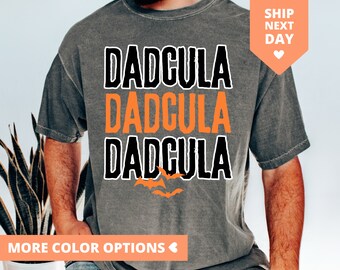 May include: A gray t-shirt with the text "Daddcula" repeated three times in black and orange. There are three orange bats flying above the text.