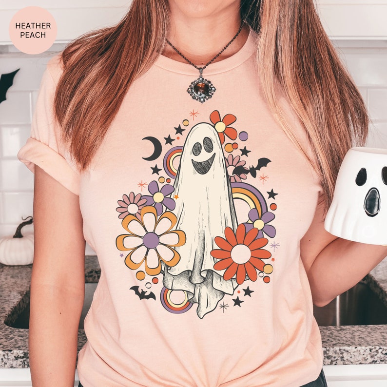 May include: A heather peach t-shirt with a retro-style graphic of a smiling ghost surrounded by flowers, stars, and a crescent moon.