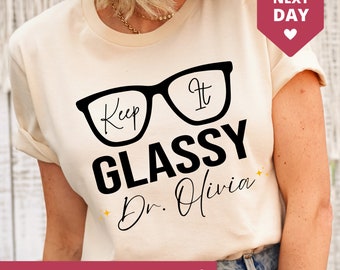 May include: A white t-shirt with a black graphic of a pair of glasses that says "Keep It Glassy" and "Dr. Olivia" underneath.