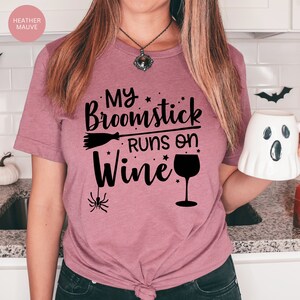 May include: A heather mauve t-shirt with the text "My broomstick runs on wine" in black. The design includes a broom, stars, and a wine glass.
