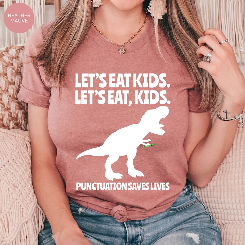 Puede incluir: Una camiseta de color malva con un gr&aacute;fico blanco de un dinosaurio comiendo un cray&oacute;n verde. El texto de la camiseta dice "LET'S EAT KIDS. LET'S EAT, KIDS. PUNCTUATION SAVES LIVES."