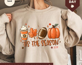Puede incluir: Una sudadera beige con un diseño gráfico que presenta una taza de café, una hoja, una pelota de fútbol, una calabaza y el texto "Tis the Season 2024".