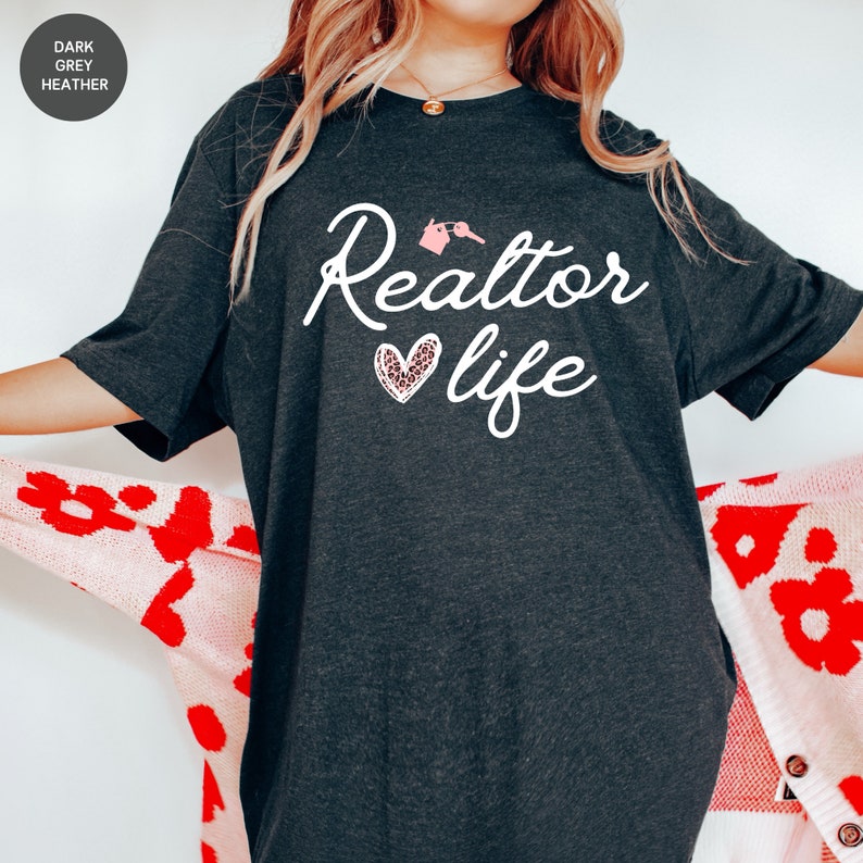 May include: A dark heather grey t-shirt with the text "Realtor life" in white. A pink heart with a leopard print design is above the word "life".