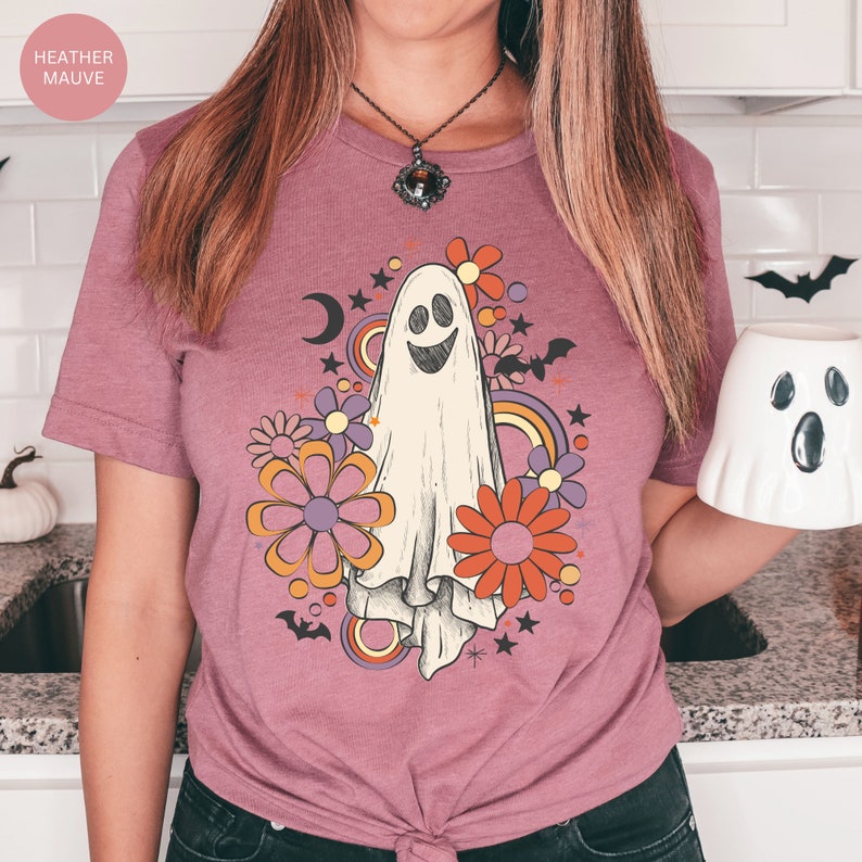 May include: A heather mauve t-shirt with a retro-style ghost design. The ghost is surrounded by colorful flowers and stars. The shirt has a short sleeve and a crew neck.