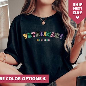 Può includere: T-shirt nero con la scritta "VETERINARY MEDICINE" in lettere colorate. Il testo è in stile collegiale. L'immagine include anche uno striscione rosa con la scritta "MORE COLOR OPTIONS".