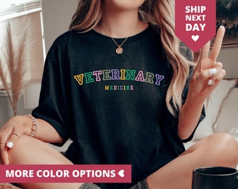 May include: Black t-shirt featuring the text "VETERINARY MEDICINE" in a vibrant, multi-coloured design. The text is in a bold, collegiate font. The image also includes a pink banner that says "MORE COLOR OPTIONS".