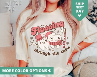 Könnte beinhalten: Cremefarbenes T-Shirt mit einem Retro-Grafikdesign von zwei Zeichentrickzähnen, einer mit Zylinder und der andere mit Weihnachtsmütze und Schal. Der Text lautet "Flossing through the snow".