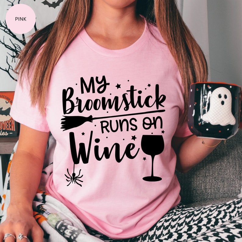 May include: A pink t-shirt with the text "My Broomstick Runs on Wine" in black. The text is accompanied by a black broom, a black wine glass, and a black spider.
