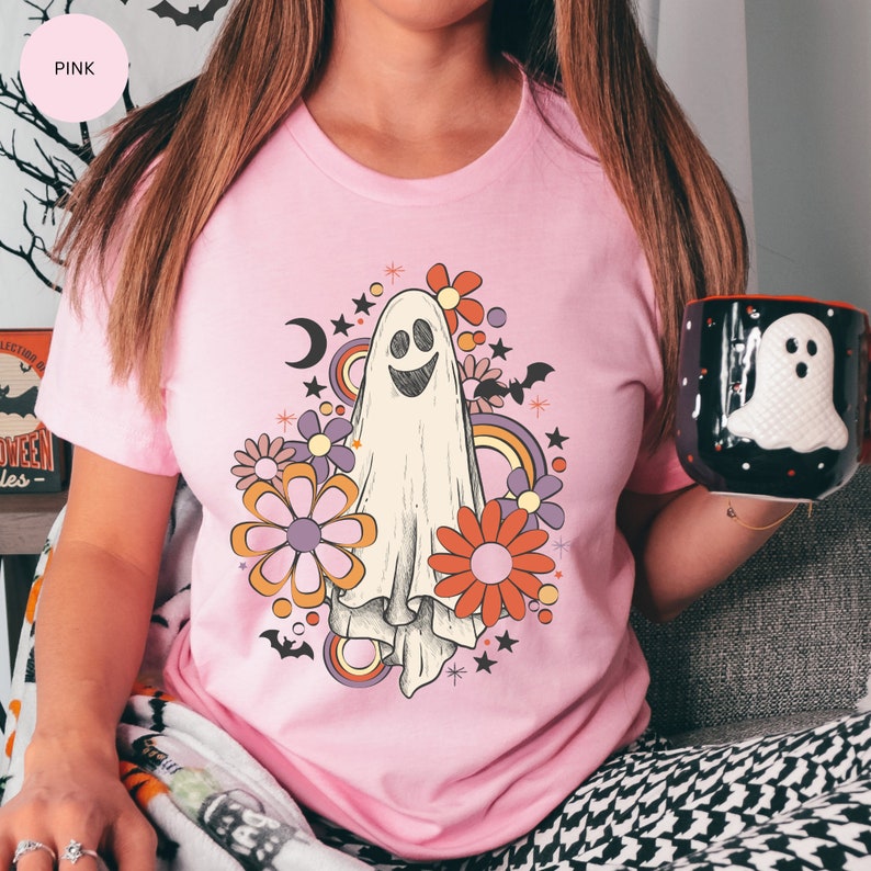 May include: Pink t-shirt with a white ghost wearing a happy face surrounded by colorful flowers and stars. The ghost is in a retro style with a rainbow and crescent moon.