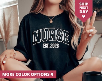 May include: Black t-shirt with the word "NURSE" in white block letters, outlined in white, above "EST. 2023". The shirt has short sleeves. The words "MORE COLOR OPTIONS" are at the bottom of the image.