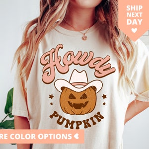 May include: A white t-shirt with a pink and brown graphic design featuring a pumpkin wearing a cowboy hat and the text "Howdy Pumpkin".