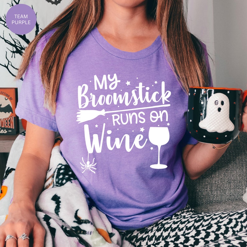 May include: A woman wearing a purple t-shirt with the text "My broomstick runs on wine" and a white graphic of a broom, stars, and a wine glass. She is holding a black mug with a white ghost design.