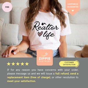 May include: White t-shirt with the text "Realtor life" in black with a pink heart and house keys graphic. The shirt is being modeled by a person sitting on a couch. The text "Happiness Guarantee" is in the top right corner of the image.