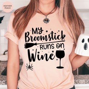 May include: A pink t-shirt with the text "My broomstick runs on wine" in black. The text is decorated with stars and a wine glass. A small black spider is hanging from the bottom of the text.