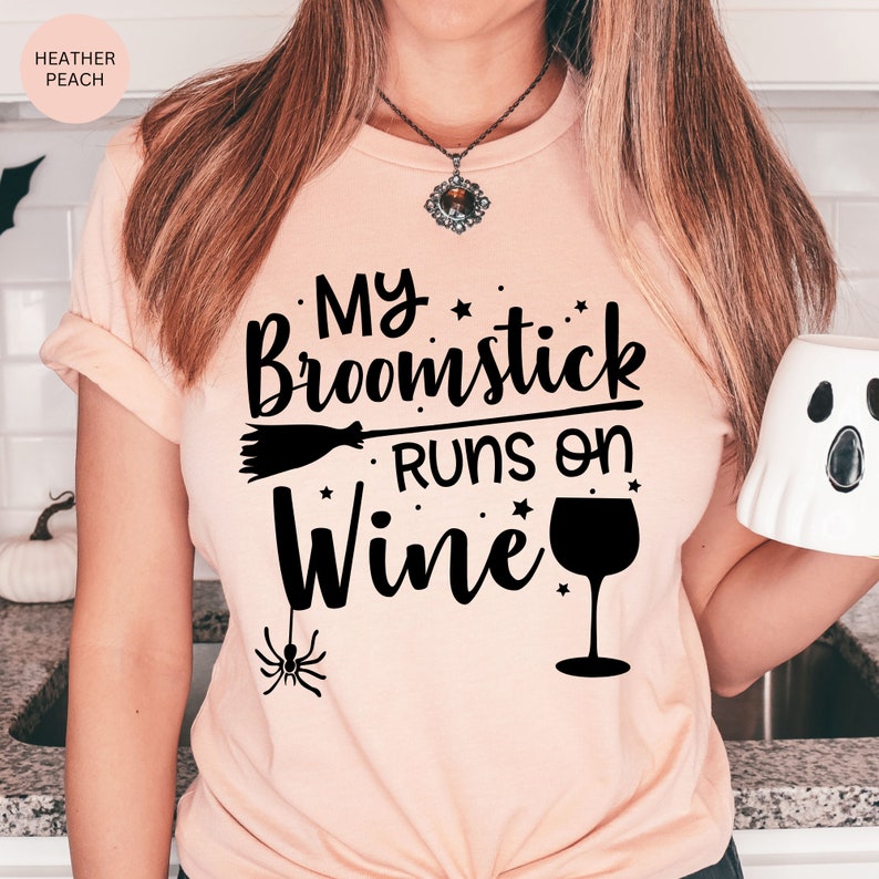 May include: A pink t-shirt with the text "My broomstick runs on wine" in black. The text is decorated with stars and a wine glass. A small black spider is hanging from the bottom of the text.