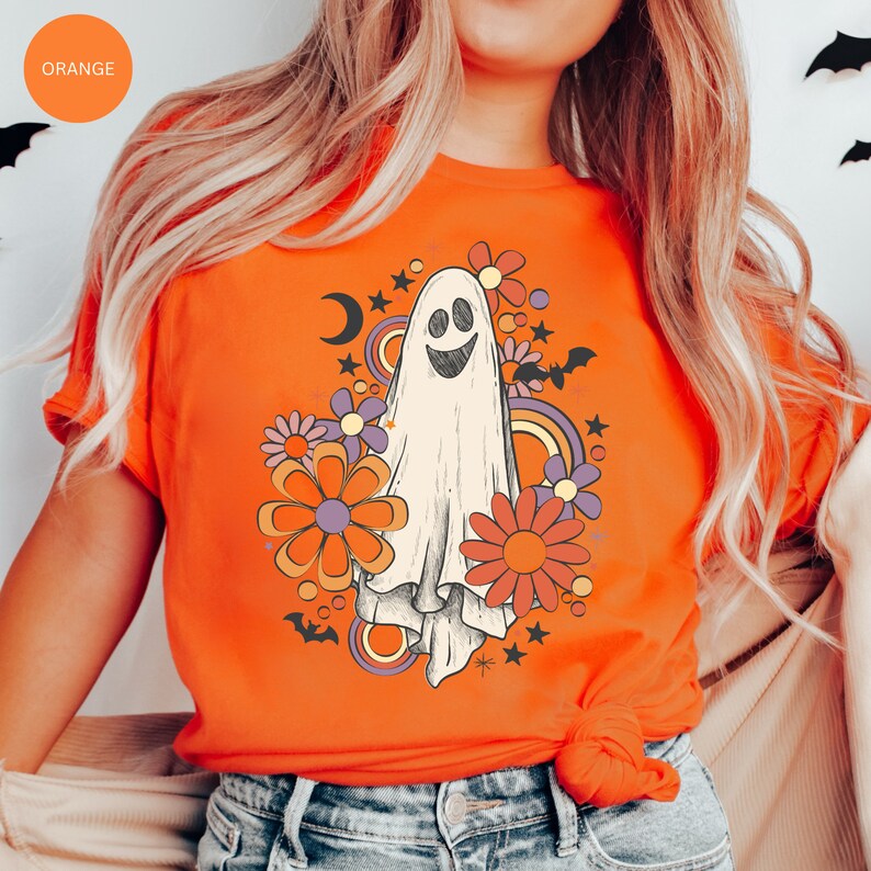 May include: Orange t-shirt with a retro-style ghost graphic. The ghost is surrounded by colorful flowers and stars. The shirt is perfect for Halloween.
